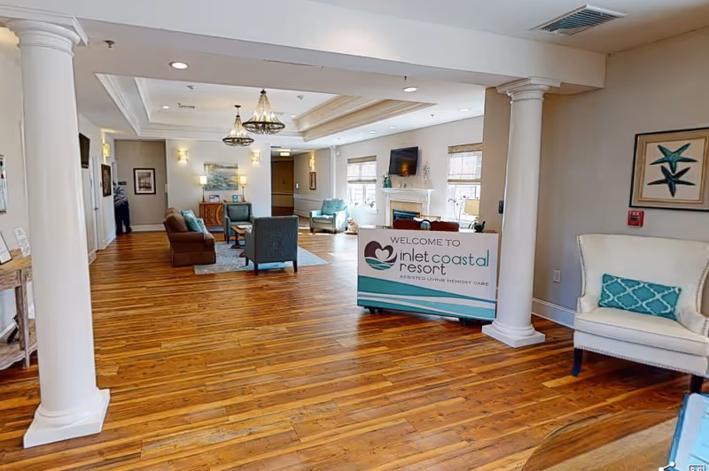 Spacious and well-lit interior common area of Inlet Coastal Resort Assisted Living and Memory Care featuring wooden floors, white columns, comfortable seating with armchairs and sofas, a fireplace with a mounted TV above it, and a welcome sign for the facility.
