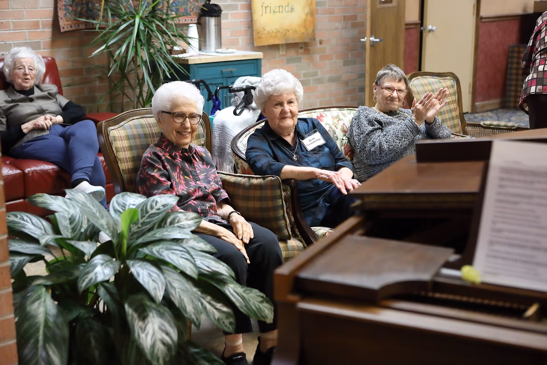 Four elderly women sitting in a cozy room with brick walls and plants. Three women are seated on cushioned chairs and a sofa, smiling and clapping, while one woman sits on a red leather couch in the background. A piano with sheet music is visible in the foreground.