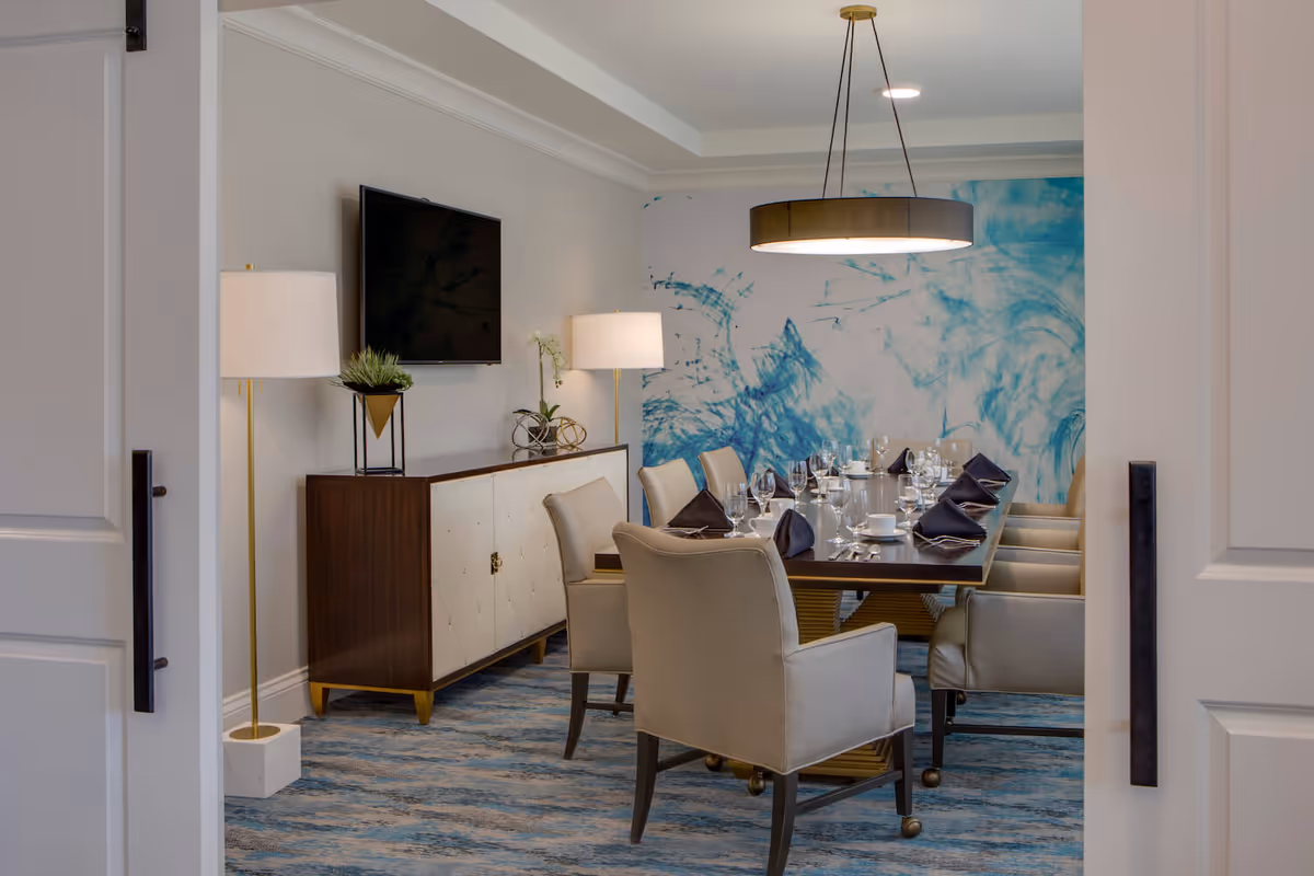 A modern dining room with a long rectangular table set for a meal, featuring beige upholstered chairs, black napkins, glassware, and white dishes. The room has a blue and white abstract wall mural, a flat-screen TV mounted on the wall, two floor lamps with white shades, and a wooden sideboard with decorative items. The floor is covered with a blue patterned carpet, and a large circular pendant light hangs above the table.