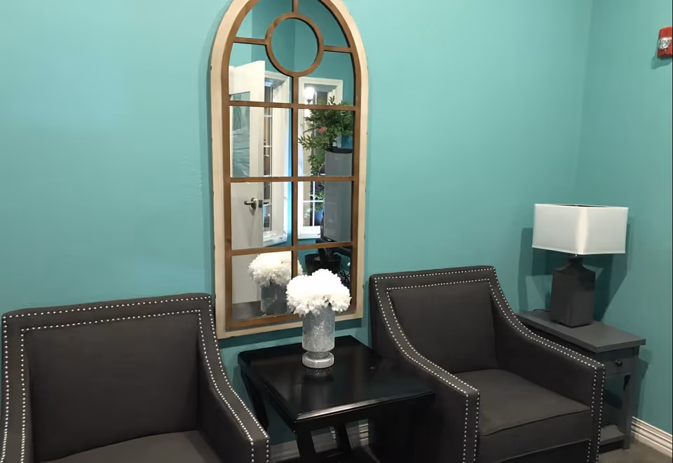 A cozy seating area with two dark gray armchairs featuring silver nailhead trim, a black side table between them holding a vase of white flowers, a gray side table with a modern lamp, and a large arched mirror with a wooden frame mounted on a teal wall.