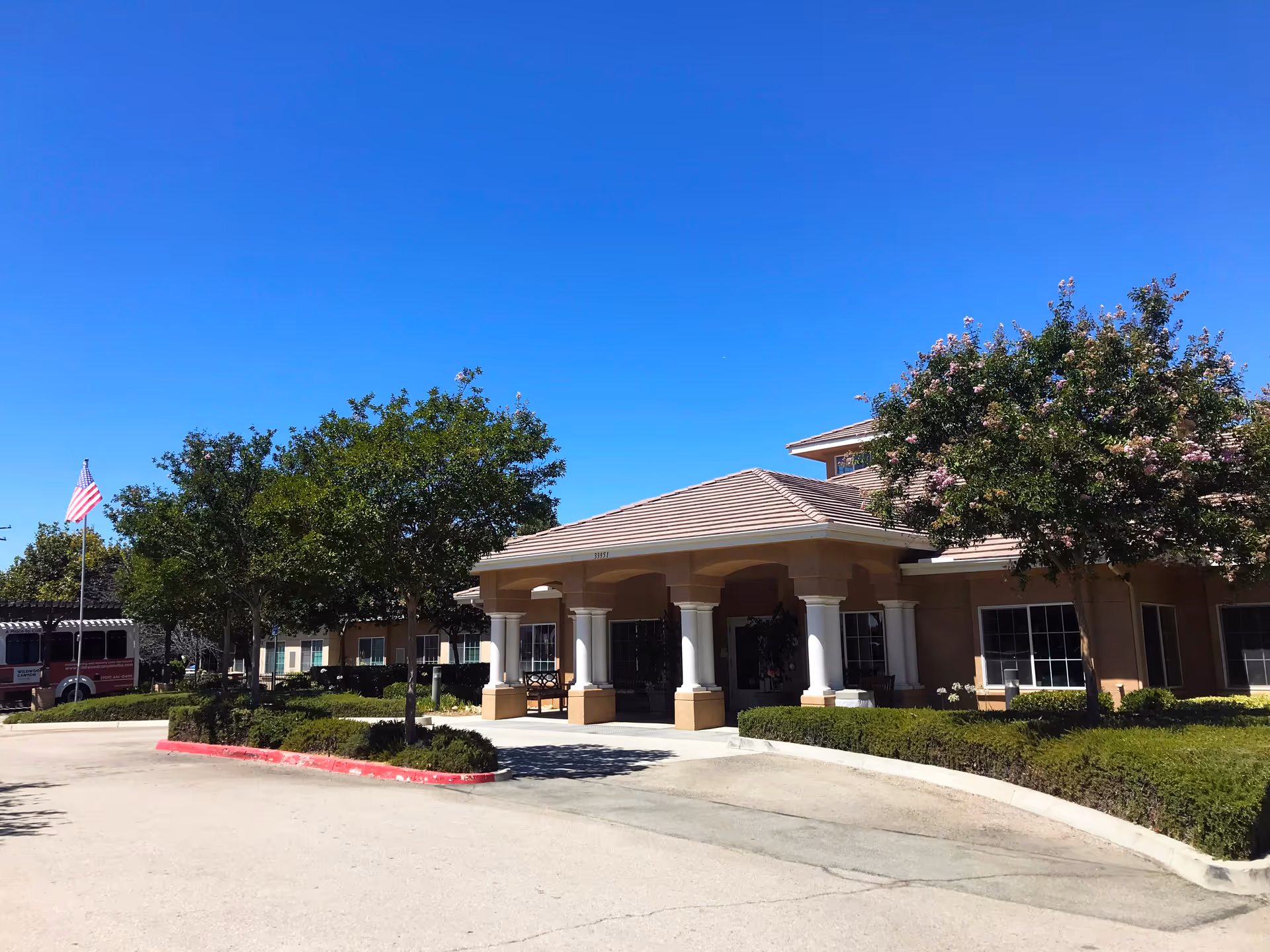 Exterior view of Wildwood Canyon Villa Assisted Living and Memory Care building with a covered entrance supported by columns, surrounded by trees and shrubs under a clear blue sky. An American flag is visible on the left side near a parked vehicle.