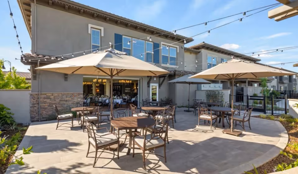 Outdoor patio area at Laurel Heights Senior Living & Memory Care with round tables and chairs under large beige umbrellas, string lights overhead, and a building with large windows in the background.