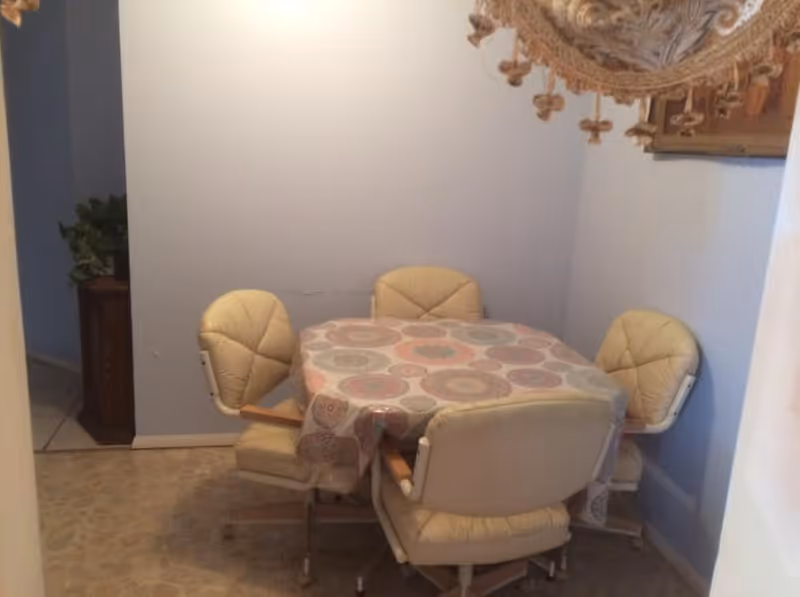 A small dining area with a square table covered by a patterned tablecloth and surrounded by four beige cushioned swivel chairs. The walls are painted light blue and there is a decorative hanging lamp above the table.