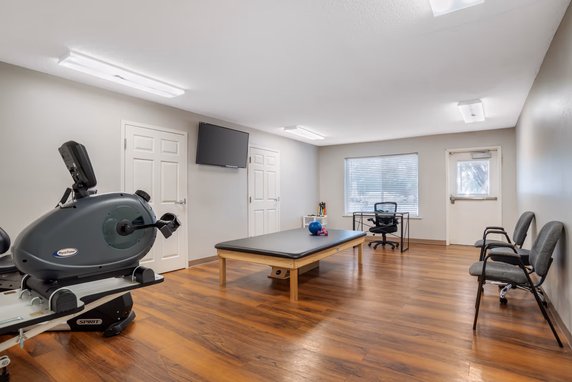 A bright interior therapy/fitness room with a recumbent exercise machine, a treatment table, desk and chairs, and a wall-mounted TV.