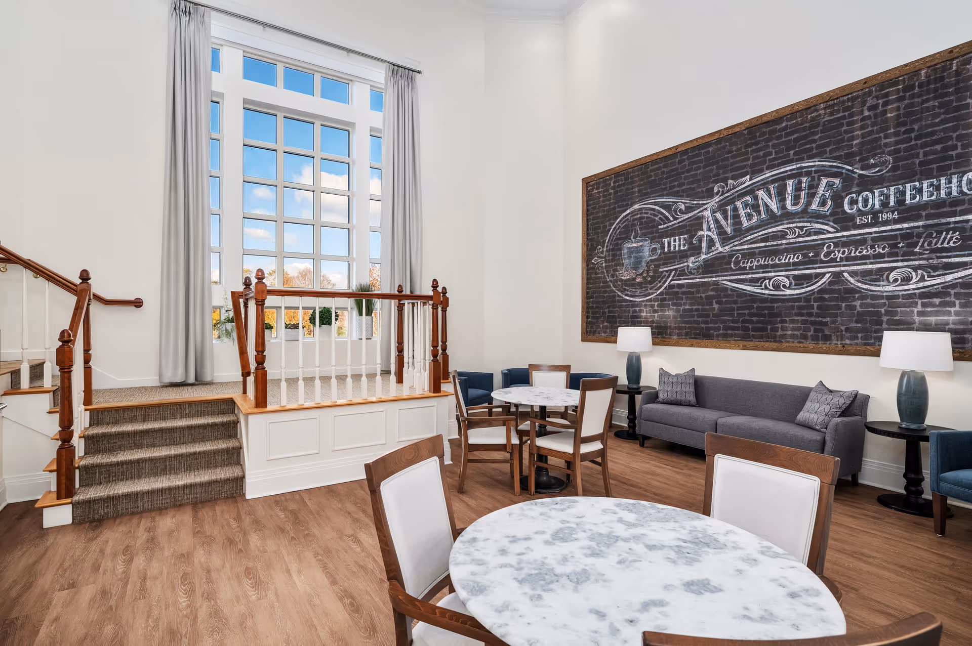 Bright communal lounge with round marble-top tables and chairs, a sofa, large window, and a chalkboard-style 'Avenue Coffeehouse' sign on the wall.