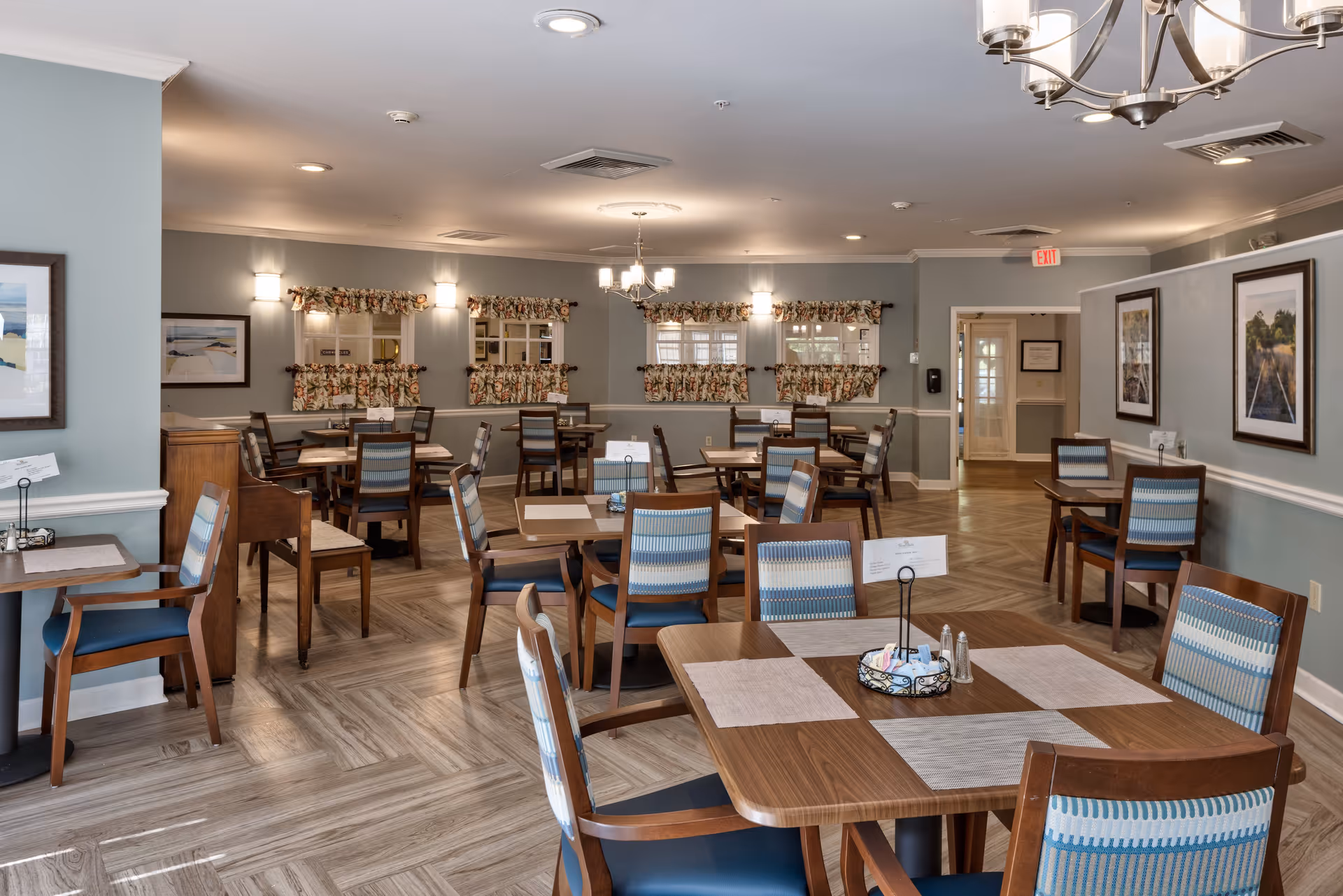 A spacious dining room with multiple wooden tables and chairs arranged neatly. The walls are painted light blue with white trim, decorated with framed artwork and floral curtains on the windows. The room is well-lit with ceiling lights and a chandelier, and the floor has a wood-patterned design.