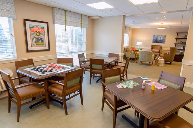 A well-lit common area in a senior living facility featuring multiple wooden tables and chairs. One table has a checkers game set up, while another table has craft supplies including glue and paper. In the background, there is a cozy seating area with armchairs, a sofa, a lamp, and a bookshelf. Large windows with blinds allow natural light to fill the room.