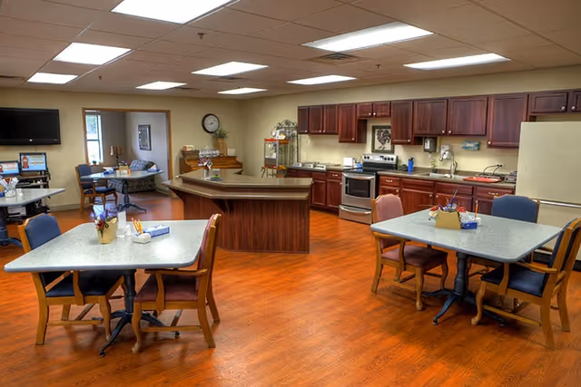 A spacious kitchen and dining area with wooden floors and cabinetry. The kitchen features stainless steel appliances including an oven and refrigerator, a large island in the center, and multiple tables with chairs arranged for dining. In the background, there is a small seating area with a TV mounted on the wall and a clock above a wooden cabinet.