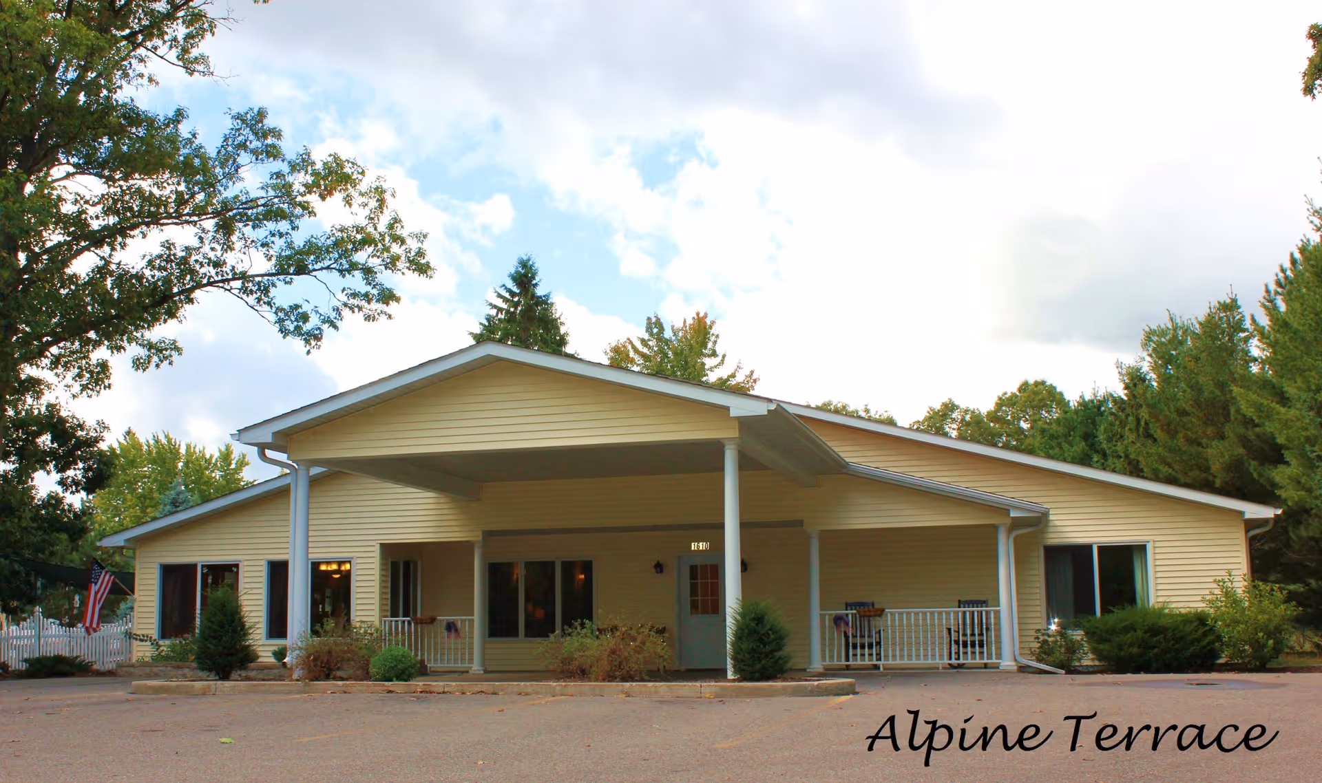 Exterior view of a single-story building with light yellow siding and a covered entrance supported by white columns. The building is surrounded by trees and bushes, with a paved area in front. The sky is partly cloudy.