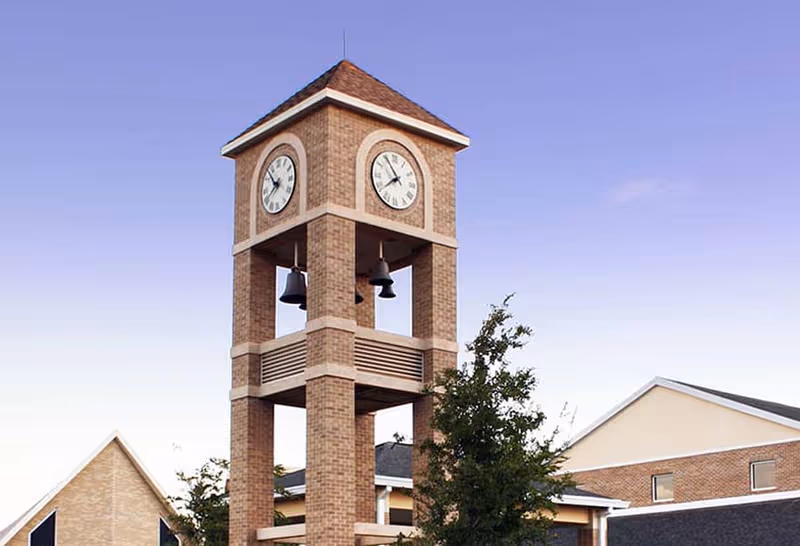 Brick clock tower with two clock faces and hanging bells beside a residential building under a clear sky.