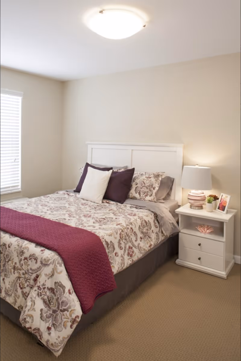 A neatly made bed with a white headboard, floral patterned bedding, and purple and white pillows in a bedroom. A purple throw blanket is draped across the foot of the bed. Next to the bed is a white nightstand with two drawers, a table lamp, a small plant, and a framed photo. The room has beige walls, a window with white blinds, and beige carpet flooring.