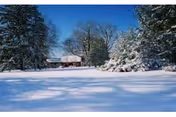 Snow-covered landscape with a small cabin or house surrounded by snow-laden trees under a clear blue sky.