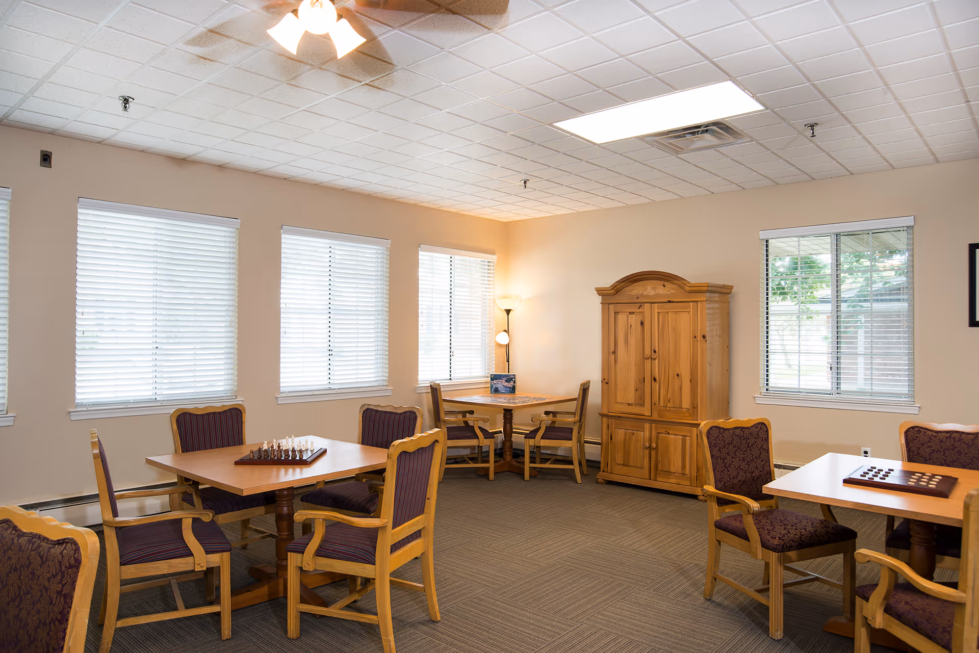 A well-lit room with multiple wooden tables and chairs arranged for games, including a chess set and a checkers board. The room has beige walls, several windows with white blinds, a wooden cabinet, and a floor lamp in the corner.