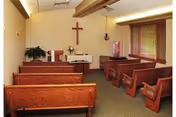 Small chapel room with wooden pews and chairs arranged facing an altar with a cross on the wall behind it. The room has beige walls, a green carpet, and windows with wooden blinds on the right side.