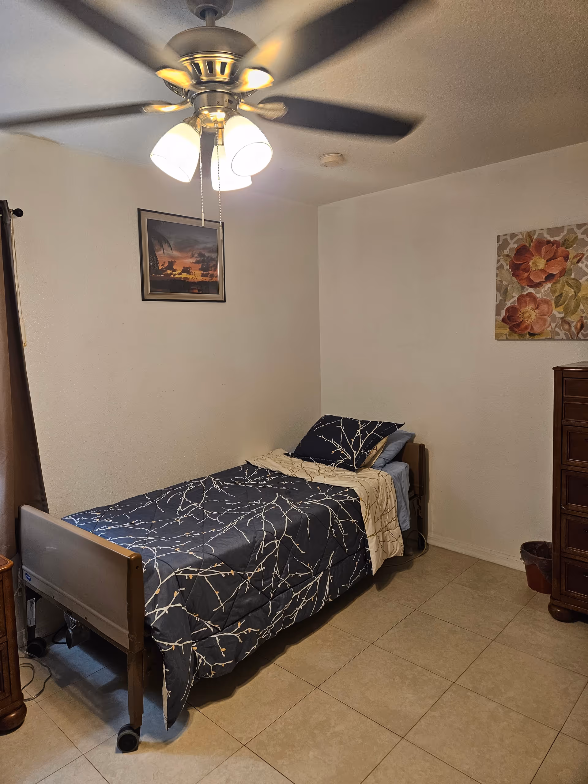 A small bedroom with a single bed on wheels, patterned bedding, a ceiling fan, and framed wall art.