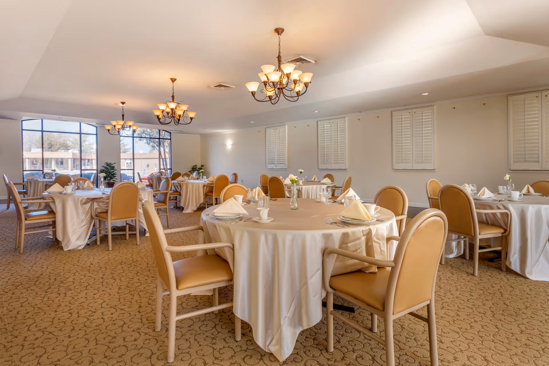A spacious dining room with round tables covered in beige tablecloths, each set with plates, cups, napkins, and glassware. The room has beige upholstered chairs around the tables, large windows letting in natural light, chandeliers hanging from the ceiling, and a carpeted floor with a subtle pattern.