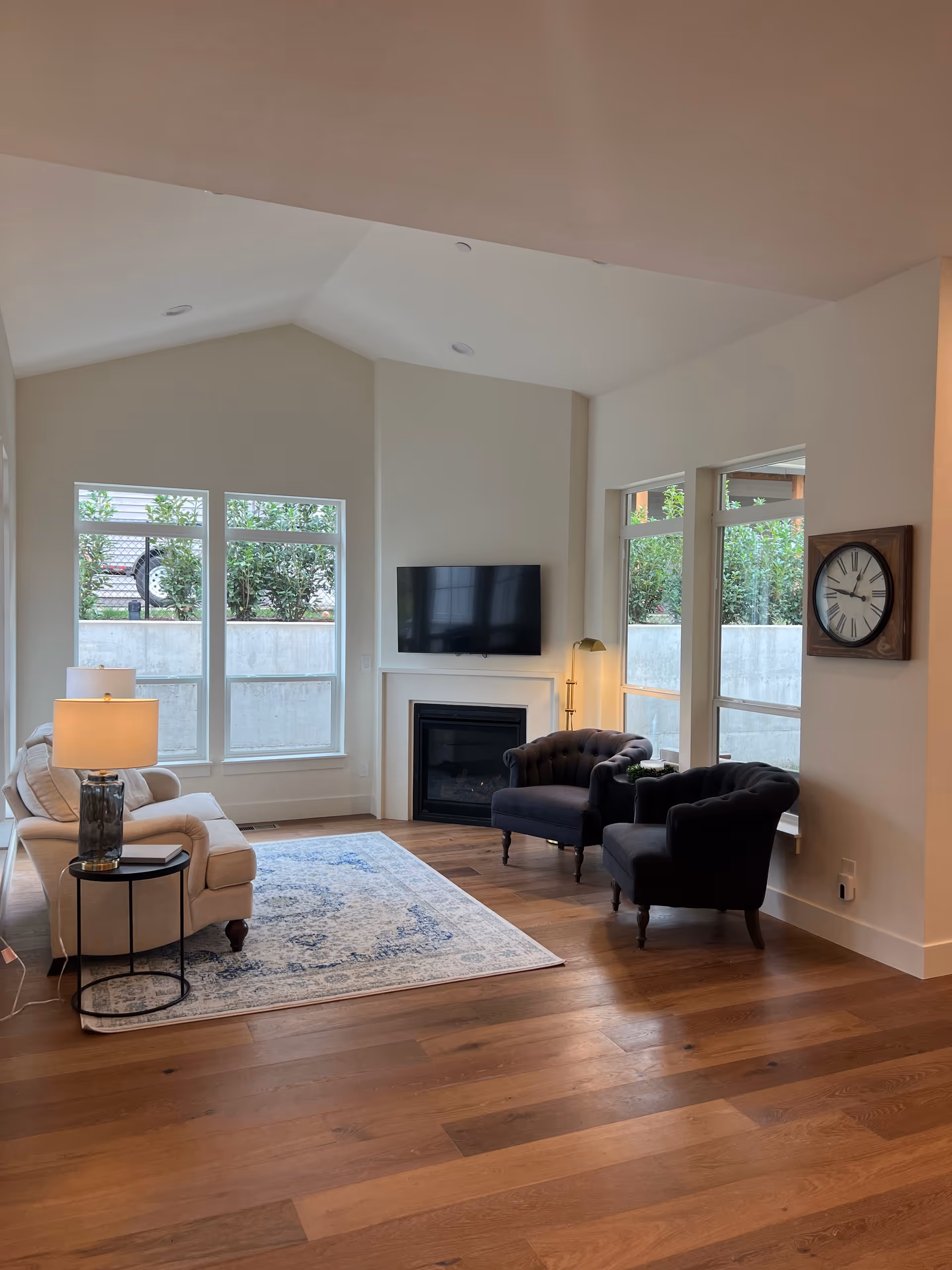 A bright living room with wooden floors, a white sofa with a side table and lamp, two dark armchairs, a wall-mounted flat-screen TV above a fireplace, large windows letting in natural light, and a wall clock.