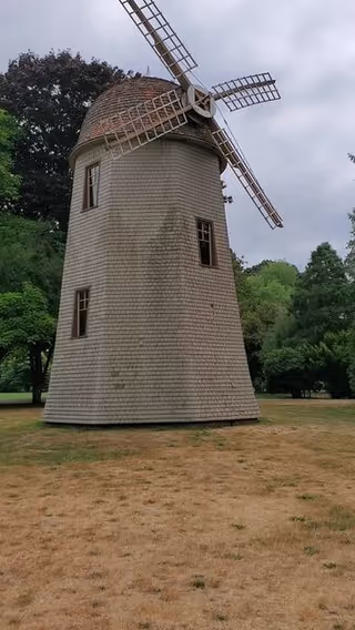 A traditional wooden windmill structure standing on a patch of dry grass with green trees and a cloudy sky in the background.