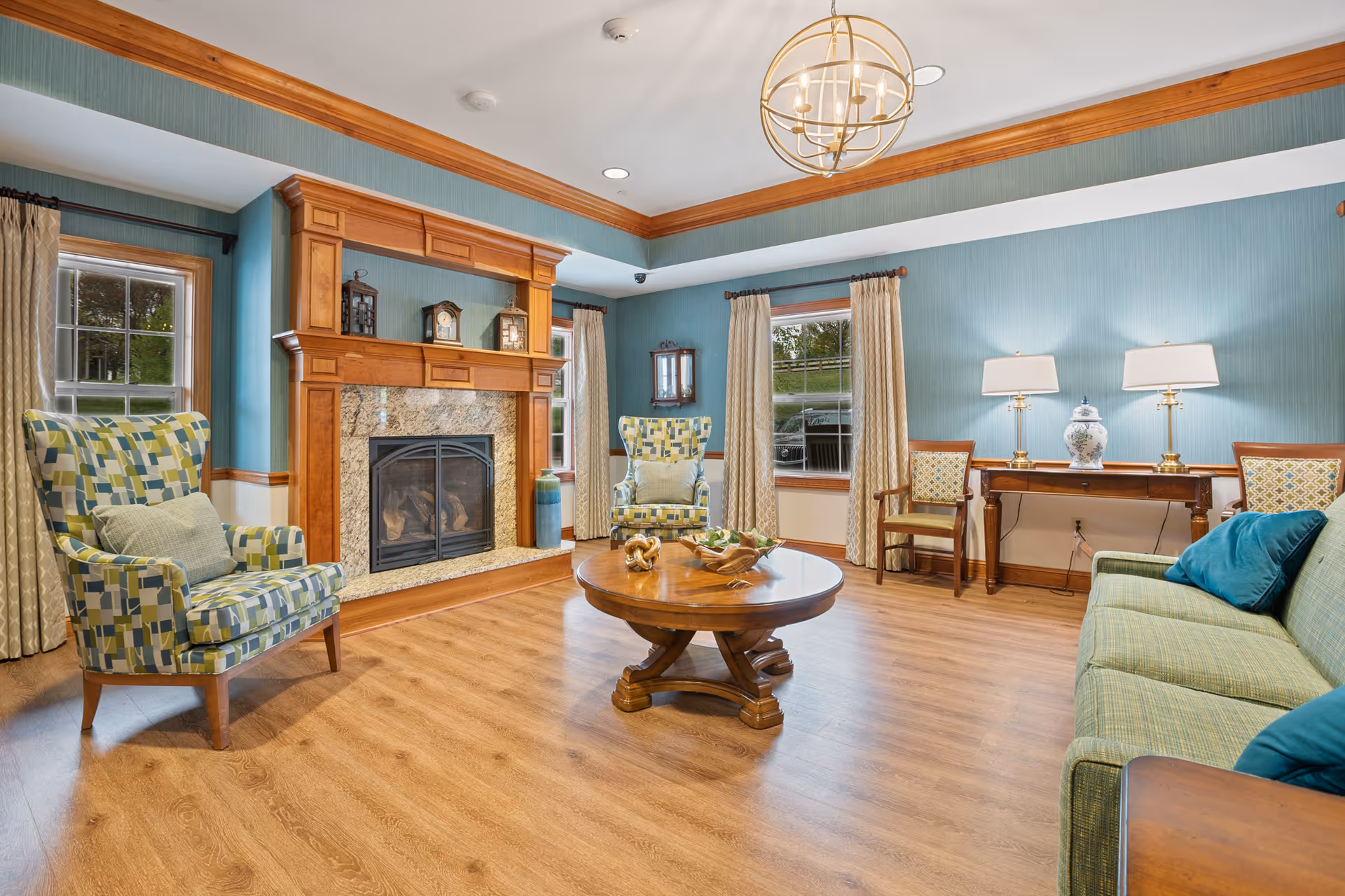 A cozy living room with wooden flooring, a wooden fireplace with a granite surround, two patterned armchairs, a green sofa with blue pillows, a round wooden coffee table with decorative items, a wooden console table with two lamps and a decorative vase, and windows with beige curtains. The walls are painted blue with wooden trim, and a modern chandelier hangs from the ceiling.