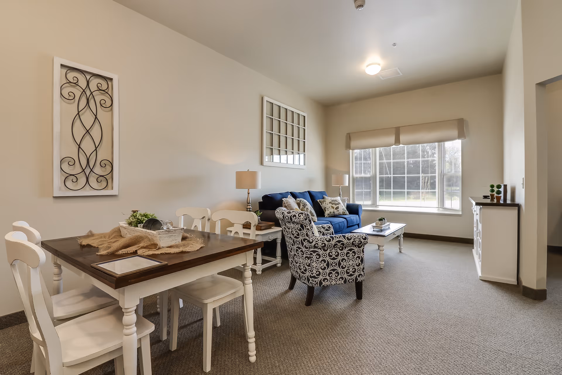 A bright and cozy living area featuring a dark blue sofa with decorative pillows, a patterned armchair, a white coffee table, and two side tables with lamps. Adjacent to the living space is a dining area with a wooden table and white chairs. The room has beige walls, a large window with blinds, and decorative wall art.