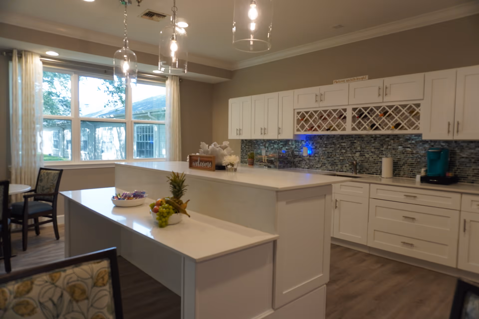A modern kitchen area with white cabinets, a mosaic tile backsplash, and a large island countertop. There are pendant lights hanging above the island, a bowl of fruit and snacks on the counter, and a coffee maker on the far right. Large windows with sheer curtains let in natural light, and there are chairs and a small table visible near the window.