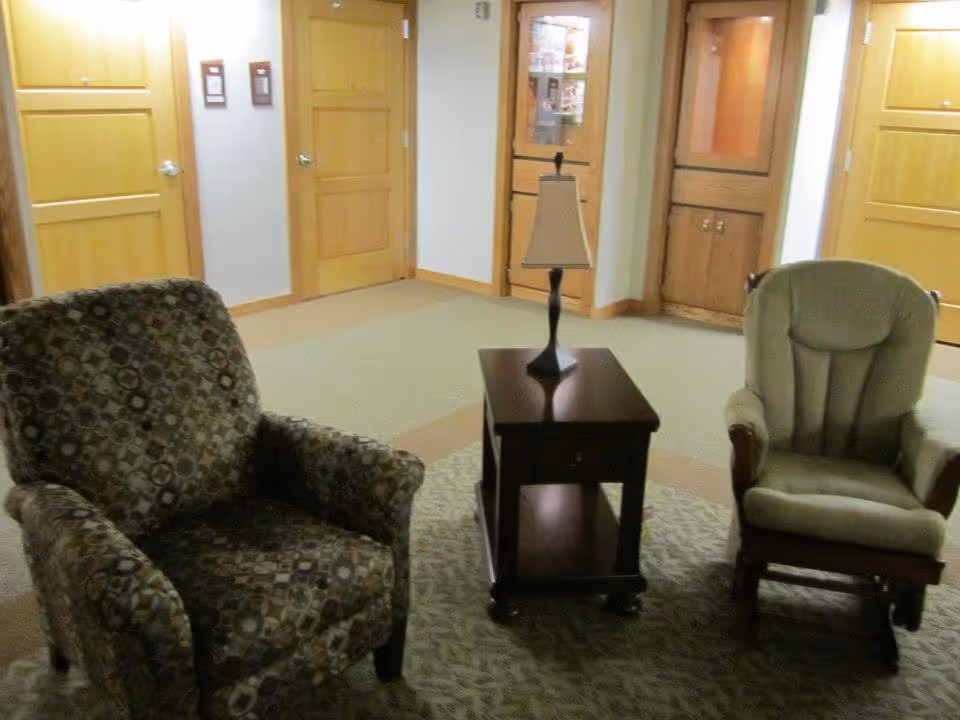 A seating area in a hallway or common space with two armchairs, one patterned and one beige, separated by a small wooden side table with a lamp on it. The background shows several closed wooden doors and a display cabinet built into the wall.