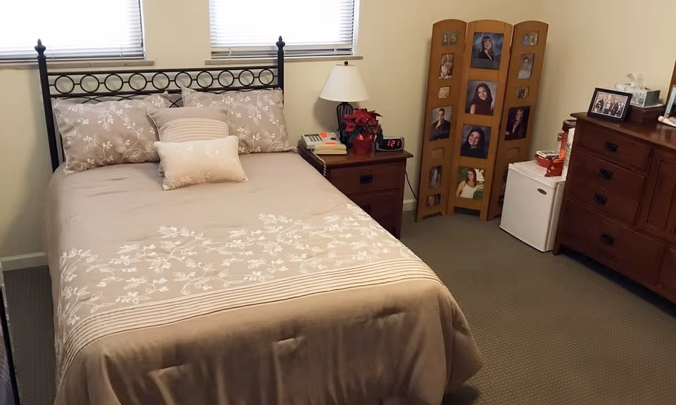 A neatly made bed with beige floral bedding and three pillows in a small bedroom. Next to the bed is a wooden nightstand with a lamp, a clock showing 1:21, a phone, and a small red poinsettia plant. In the corner, there is a wooden photo frame stand displaying multiple portraits. A small white mini-fridge is beside a wooden dresser with several drawers, topped with framed photos and tissue box. The room has two windows with blinds partially closed.