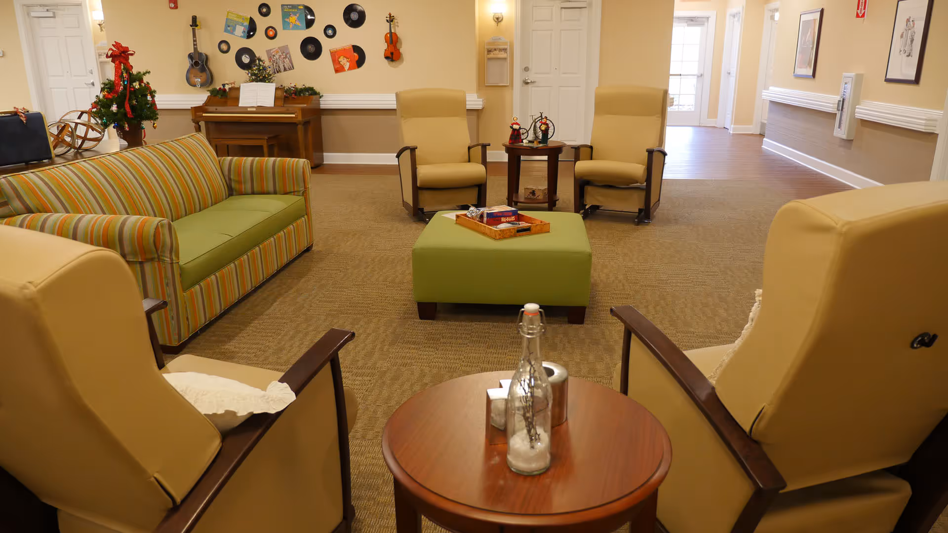 A cozy common area in a senior living facility with four beige armchairs arranged around a small wooden table and a green ottoman in the center. There is a striped green and orange couch on the left side. The background features a piano with a small decorated Christmas tree on top, wall-mounted guitars, vinyl records, and framed pictures. The room has beige walls and carpeted flooring with a hallway leading to other rooms.