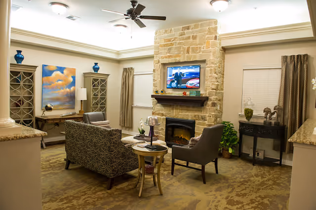A cozy living room area with a stone fireplace and a mounted flat-screen TV above it. The room features a patterned sofa, two armchairs, a small round wooden table, and decorative cabinets with blue vases on top. There are two windows with blinds and curtains, a ceiling fan, and warm lighting creating a comfortable atmosphere.