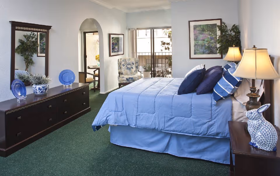 A bedroom with a neatly made bed covered in light blue bedding and multiple pillows. There is a wooden nightstand with a lamp and a decorative blue and white ceramic rabbit. Across from the bed is a dark wooden dresser with a large mirror above it, decorated with blue plates and a plant. The room has green carpet, a floral armchair near a sliding glass door leading to a balcony, and framed artwork on the walls.