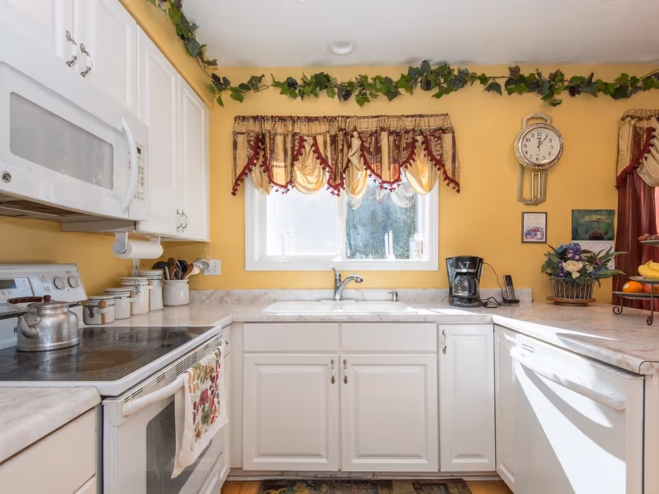 A bright kitchen with white cabinets and countertops, a stove with a kettle, a microwave, a coffee maker, and a window with decorative curtains. There is a wall clock and some decorative plants and flowers on the counter.