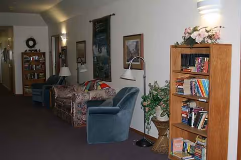 A cozy senior living lounge with teal chairs, sofa, lamps, and a bookshelf.