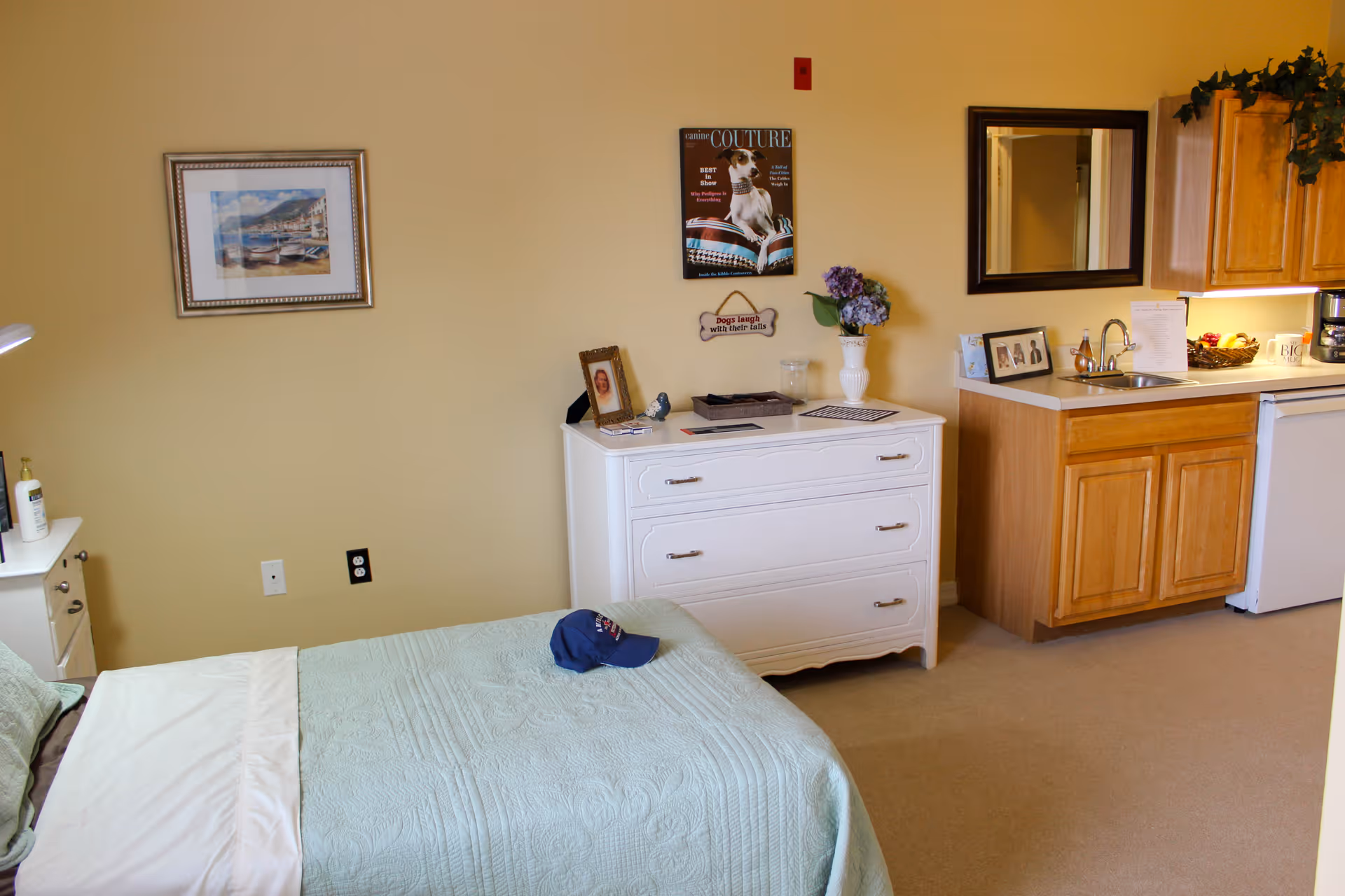 A cozy room featuring a single bed with a light green quilt and a blue cap on it. Next to the bed is a white dresser with framed photos, a vase with flowers, and decorative items. On the wall above the dresser is a framed picture and a poster with a dog. To the right is a small kitchenette area with wooden cabinets, a sink, a mini fridge, and a mirror above the counter. The walls are painted light yellow and the floor is carpeted.