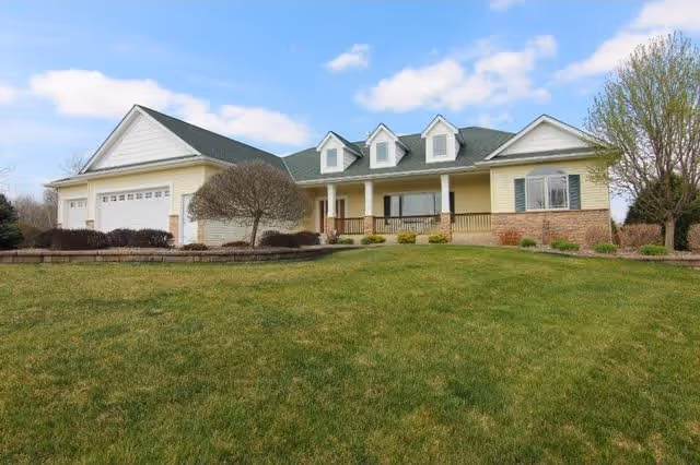 Single-story house with a green roof, three dormer windows, a front porch with railings, and a large green lawn in front under a partly cloudy sky.