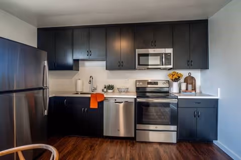 Modern kitchen with dark cabinets, stainless steel refrigerator, dishwasher, oven, and microwave. The countertop has a paper towel holder, a small plant, a bowl, a vase with yellow flowers, and a cutting board. The floor is wooden.