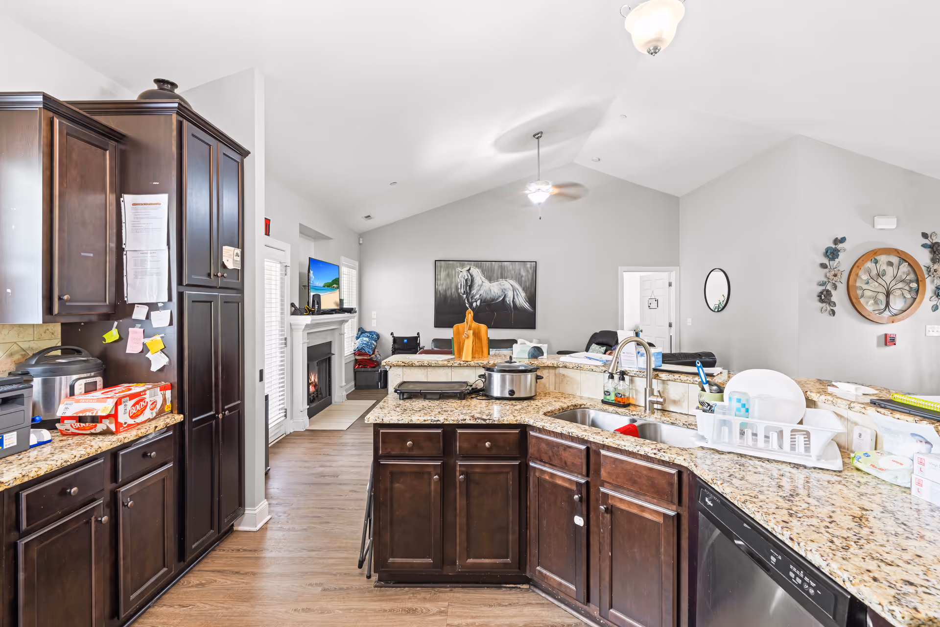 Open kitchen with dark wood cabinets and granite countertops looking into a living room with a fireplace.