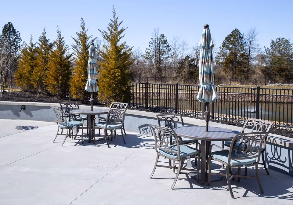 Outdoor patio area with two round tables, each surrounded by four metal chairs with blue cushions. Each table has a closed striped umbrella in the center. The patio is paved with concrete and bordered by a low wall and a black metal fence. Behind the fence, there are trees and a small body of water under a clear blue sky.