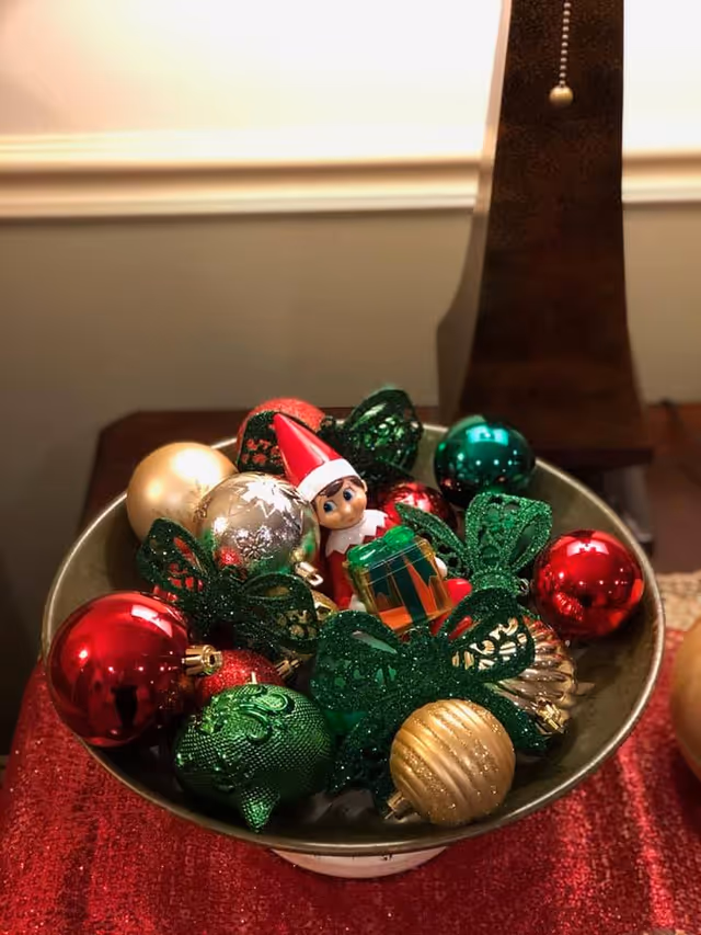 A decorative bowl filled with colorful Christmas ornaments including red, green, gold, and silver baubles, green glittery bows, and a small elf doll holding a wrapped gift, placed on a red cloth on a wooden surface with a lamp in the background.