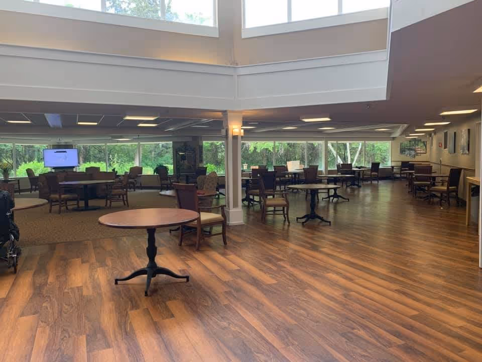 Spacious senior living dining/activity room with scattered round tables and chairs and large windows overlooking trees.