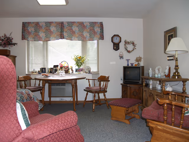 A cozy living room with vintage wooden furniture including armchairs, a small table with two chairs, a TV on a wooden cabinet, and decorative items such as a wall clock, floral arrangements, and framed pictures on the window sill. The room has a carpeted floor and a window with floral valance curtains.