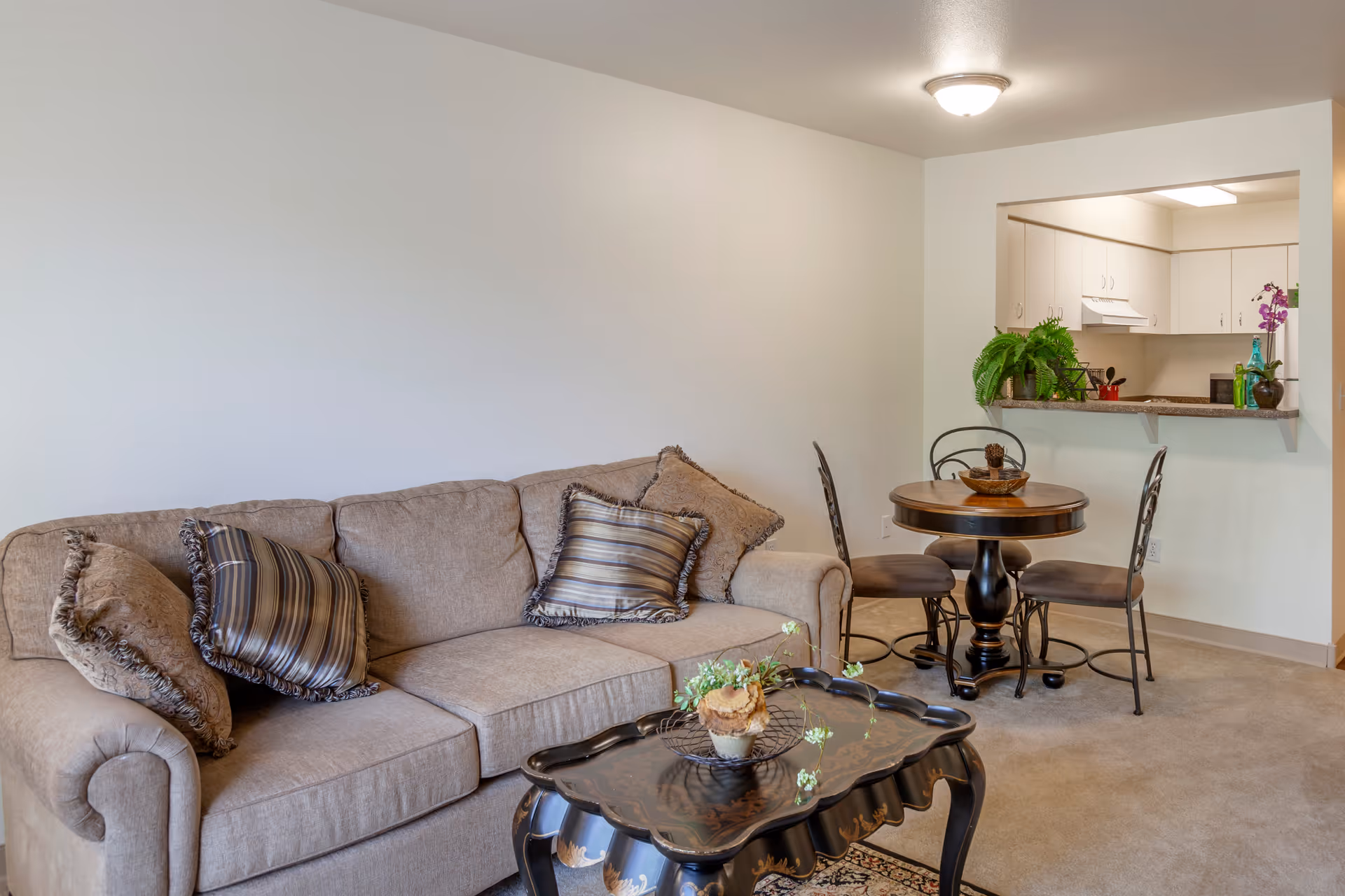 A cozy living area featuring a beige sofa with four decorative pillows, a black ornate coffee table with a small plant centerpiece, and a round wooden dining table with three chairs. The dining area is adjacent to a kitchen with white cabinets and a pass-through window. The room is carpeted and has neutral-colored walls with a ceiling light fixture.