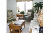 A cozy living room with a floral patterned sofa, two green armchairs, a wooden coffee table with magazines and a flower vase, a side table with a lamp and a plant, and a large window with sheer curtains letting in natural light.
