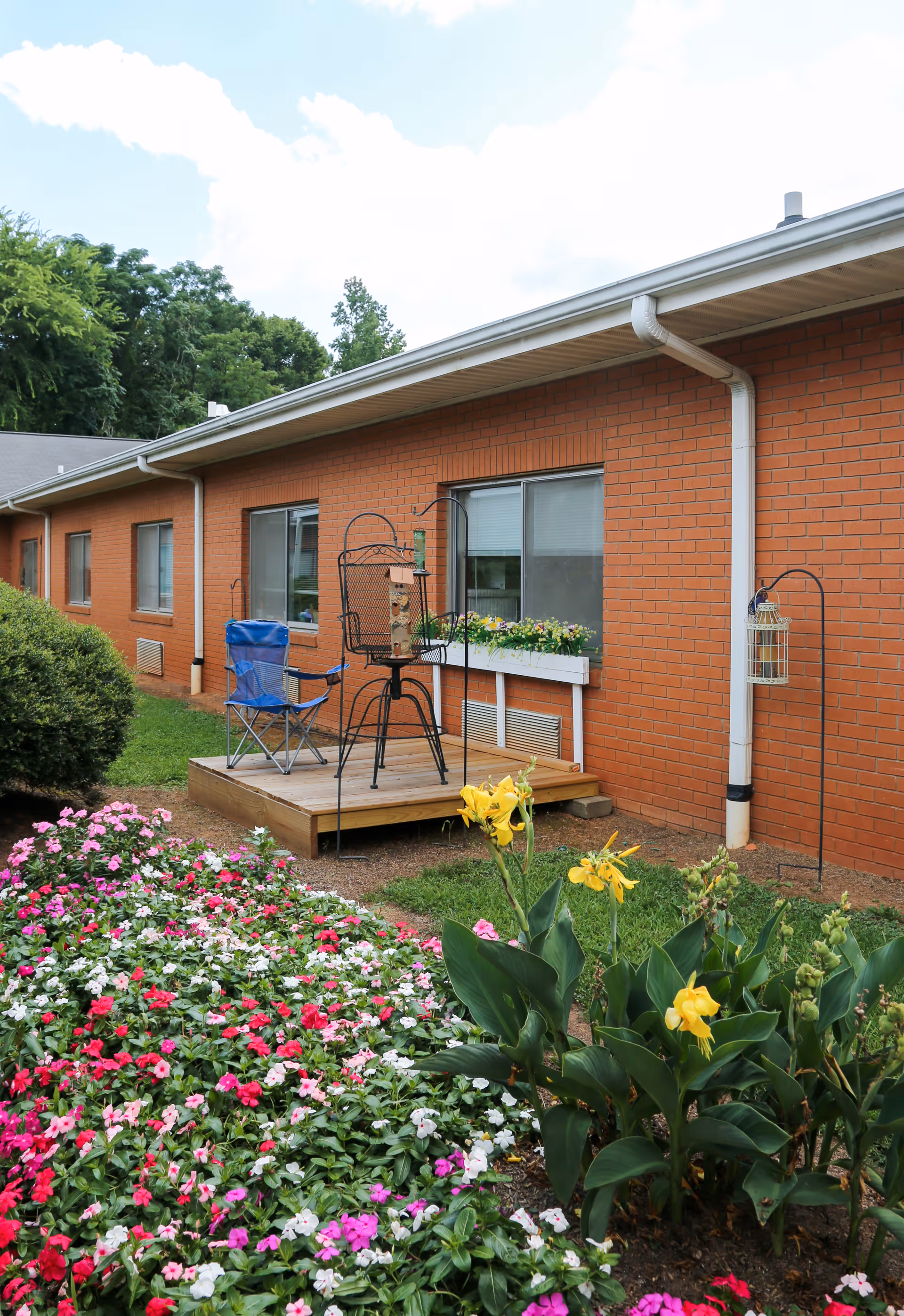 Brick exterior of a single-story senior facility with a small wooden deck, folding chair, window flower box and colorful flower beds in front.