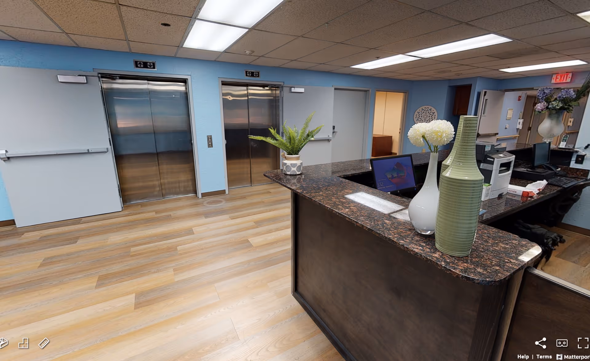 Interior view of a facility lobby area with two stainless steel elevators on a light blue wall. In the foreground, there is a dark granite countertop reception desk with decorative vases and flowers. The floor is wood-patterned, and there is an exit door and office area visible in the background.