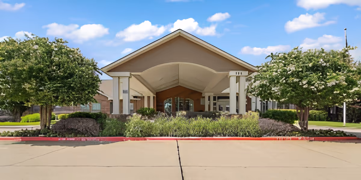 Front exterior view of Morada Cedar Hill facility with a covered entrance, flanked by two trees and landscaped bushes under a blue sky with scattered clouds.