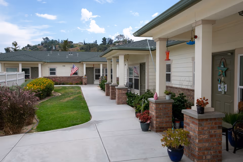 Outdoor walkway at Lo-Har Senior Living with a concrete path bordered by green grass and plants. The single-story building has a covered porch area with brick pillars and several potted plants. An American flag is displayed near one of the doors, and the sky is partly cloudy.