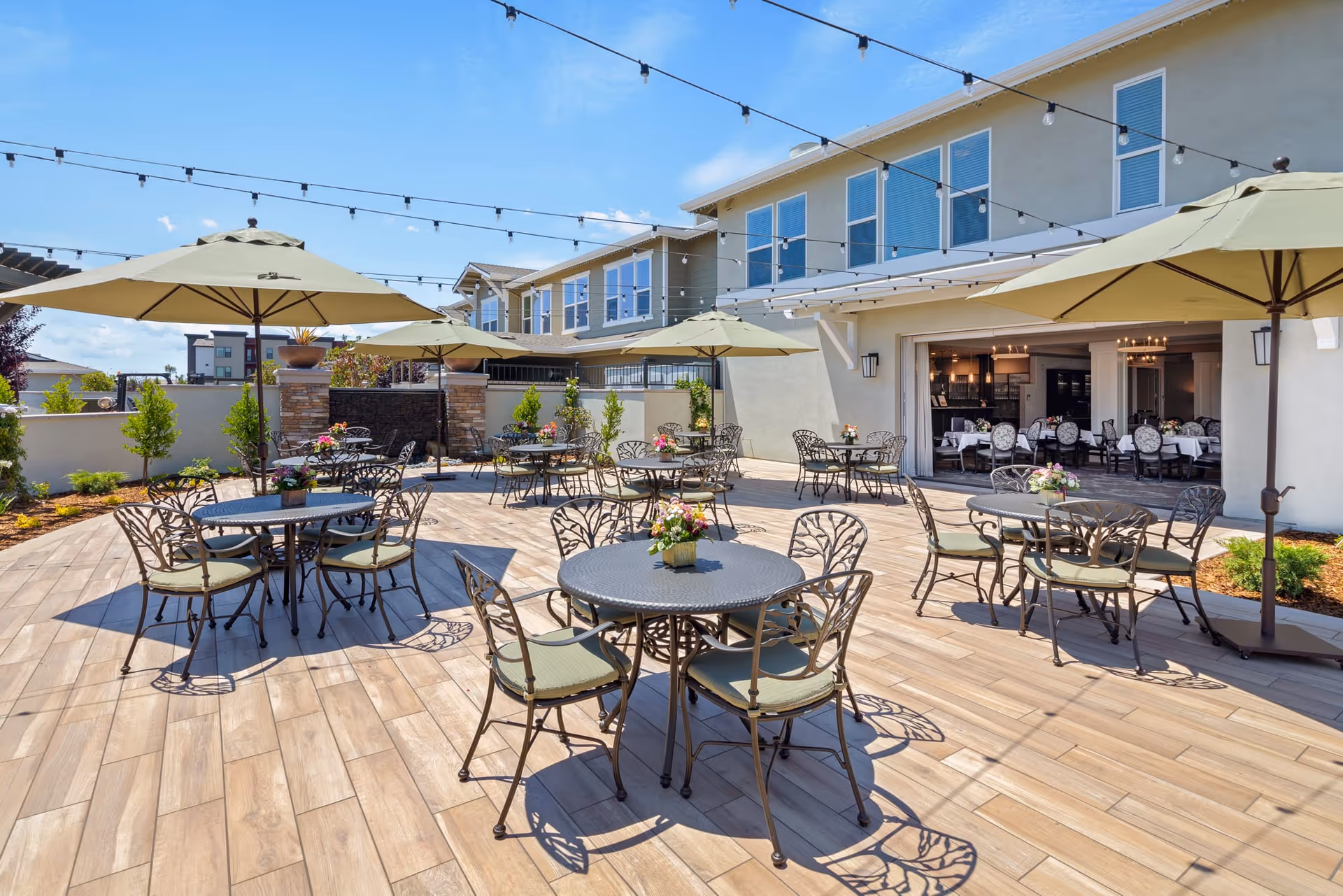 Outdoor patio area at Oakmont of Lodi with multiple round metal tables and chairs, each table having a small flower arrangement. Large green umbrellas provide shade over some tables. String lights are hung above the patio. The patio is adjacent to a building with large windows and an open dining area visible inside.