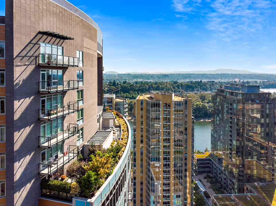 High-rise residential building with balconies and a rooftop garden overlooking a river and neighboring towers under a clear blue sky.
