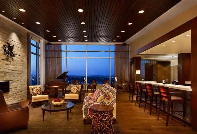 A spacious and elegantly furnished living room with large floor-to-ceiling windows offering a city view at dusk. The room features a grand piano, patterned sofas and armchairs, a round coffee table, a bar area with high stools, and warm lighting with a wooden ceiling and stone accent wall.