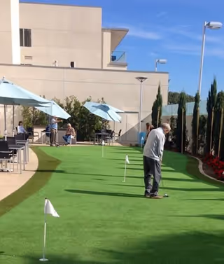 An outdoor putting green at a senior living facility with several small flags marking holes. A man is putting on the green while other people sit under blue umbrellas at tables nearby. The area is surrounded by buildings, trees, and a clear blue sky.